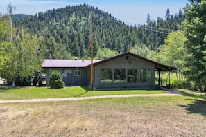 A two-story building with a metal roof and large windows, surrounded by a forested area and a clear sky.