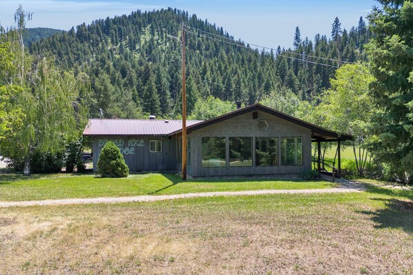 A two-story building with a metal roof and large windows, surrounded by a forested area and a clear sky.