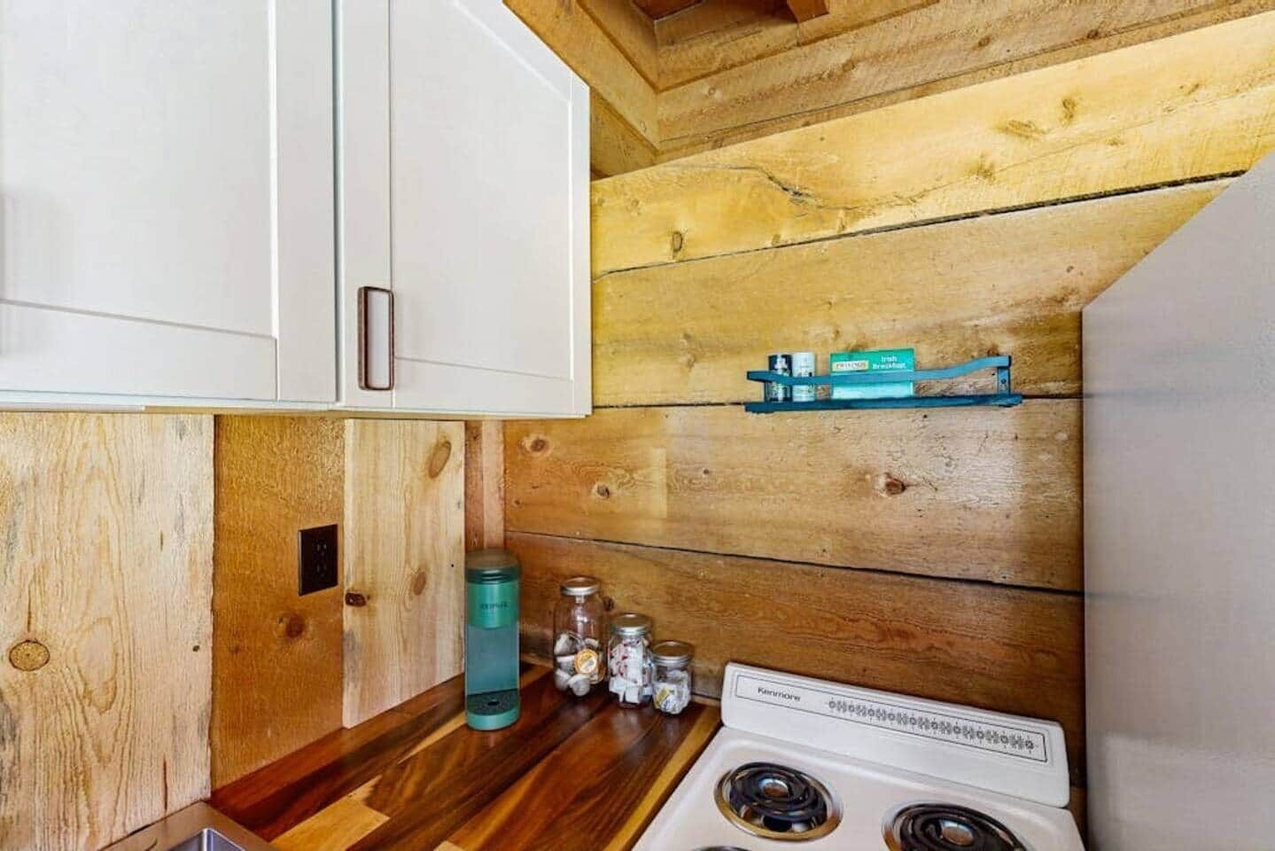Cocina pequeña con paredes de madera, una estufa blanca y un estante con utensilios de cocina.