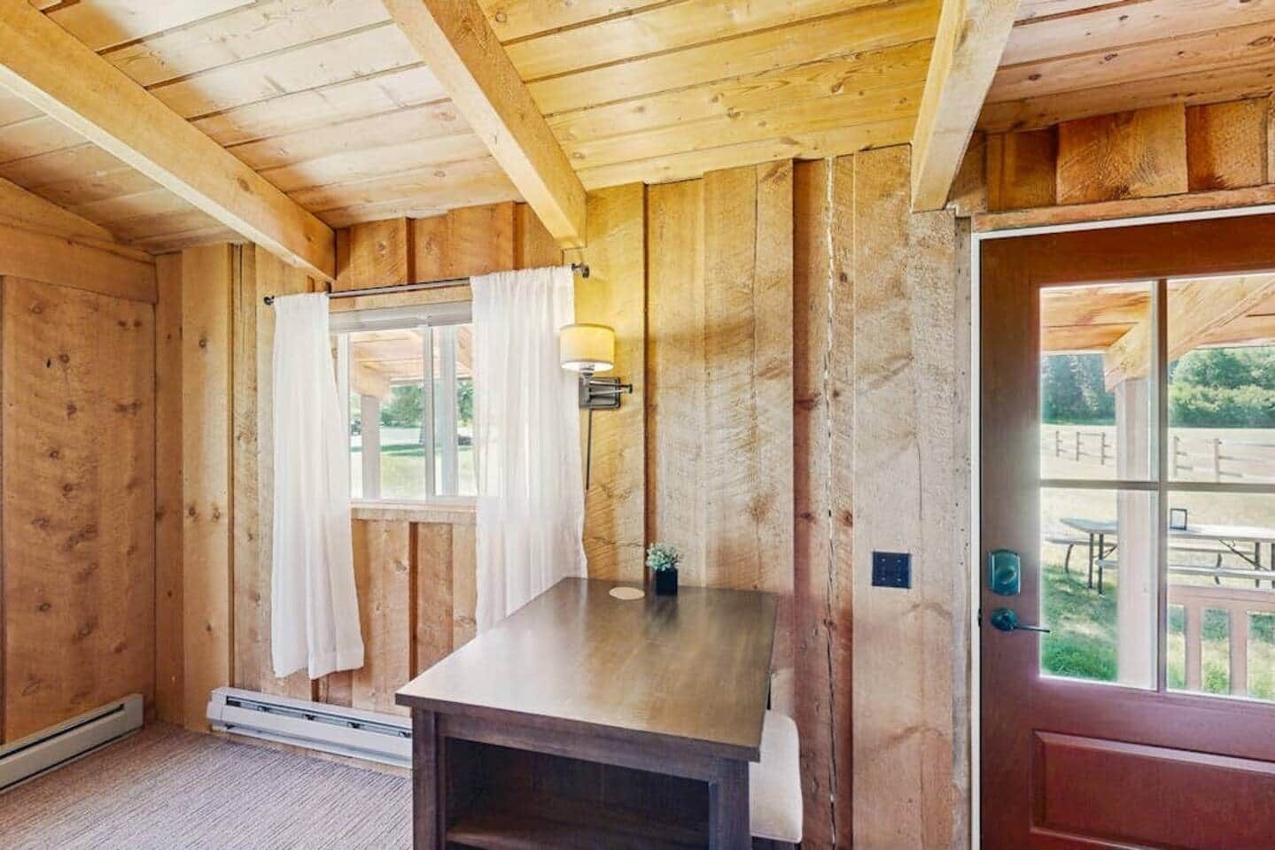 Interior de una cabaña de madera con una mesa, una ventana con cortinas y una puerta que da al exterior.
