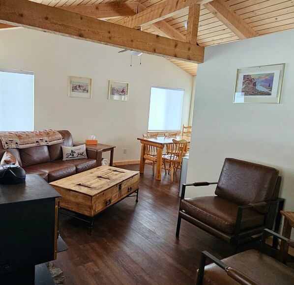 A spacious living room with a wooden ceiling, a brown leather sofa, a wooden coffee table, and a dining area with a wooden table and chairs.