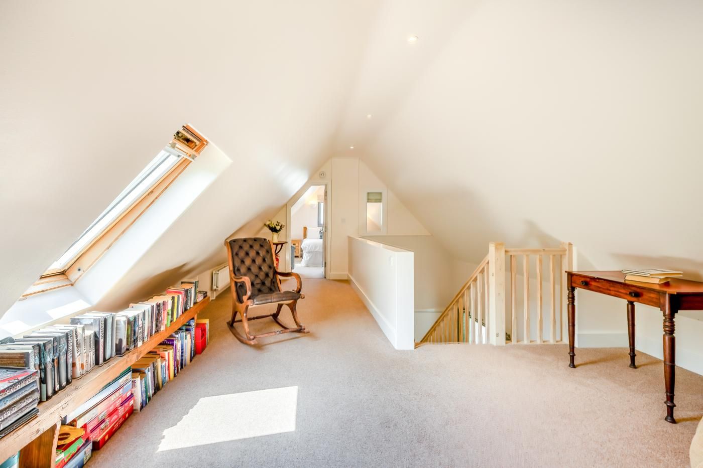 Une pièce mansardée spacieuse, avec un plafond en pente, un fauteuil à bascule en bois, une bibliothèque remplie de livres et une petite table en bois.