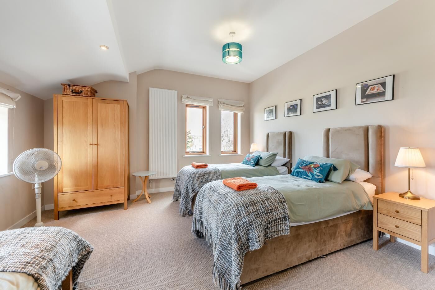 A bedroom with two beds, a wooden wardrobe, a standing fan, and a window with blinds.