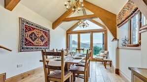 A dining area with a wooden table set for four, a chandelier, and a large window offering a view of the outdoors. - 10 Bed in Howtown (oc-w33505) (Ullswater)