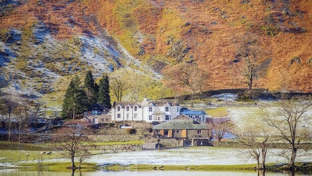 A large, white, multi-story house with a stone foundation, situated by a calm lake with reflections of the surrounding autumn-colored trees. - 10 Bed in Howtown (oc-w33505) (Ullswater)