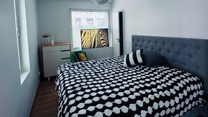 Une chambre moderne avec un grand lit, une tête de lit rembourrée de couleur grise et une fenêtre avec des stores.