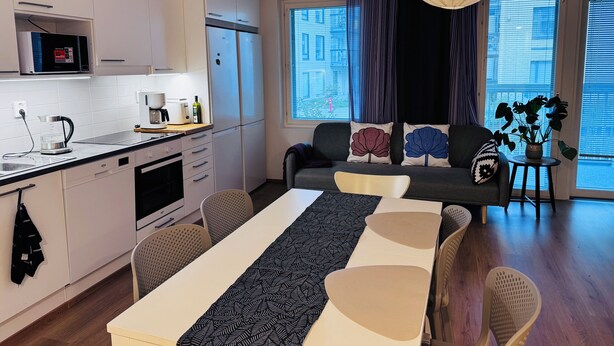 Une cuisine moderne avec des meubles blancs, un four à micro-ondes et une machine à café. On y trouve un coin repas avec une table et des chaises, ainsi qu’un canapé agrémenté de coussins décoratifs. Une grande fenêtre avec des rideaux est visible, et un luminaire suspendu est également présent.
