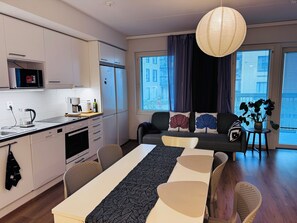 A modern kitchen with white cabinets, a microwave, and a coffee maker. There is a dining area with a table and chairs, and a sofa with decorative pillows. A large window with curtains is visible, and a hanging light fixture is present. - Nordic Home Bliss 2BR (Helsinki)