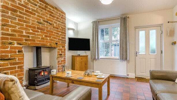 A living room with a brick fireplace, a television mounted on the wall, a window with curtains, and a door. - 3 Bed in Wells-next-the-Sea (oc-h33452) (Wells-next-the-Sea)