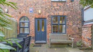 A brick building with a dark blue door, two windows, a bench, and a table. - 3 Bed in Wells-next-the-Sea (oc-h33452) (Wells-next-the-Sea)