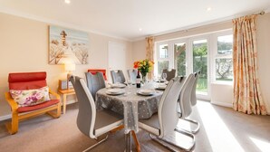 A dining room with a round table, chairs, a painting on the wall, and a window with curtains. - 4 Bed in Shaldon (oc-sunnhs) (Shaldon)