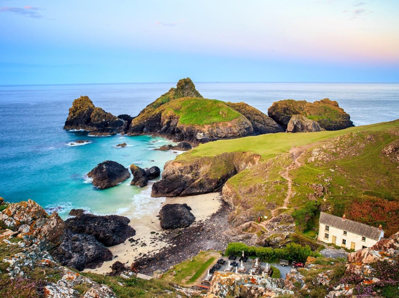 A coastal landscape with rocky outcrops, a grassy hill, and a white building with a path leading to it.