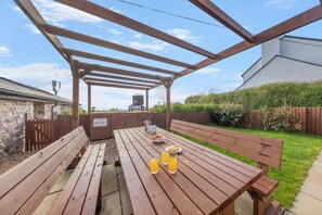 Ulkokäyttöön tarkoitettu puinen pergola, jossa on grilli, piknikpöytä ja penkkejä.