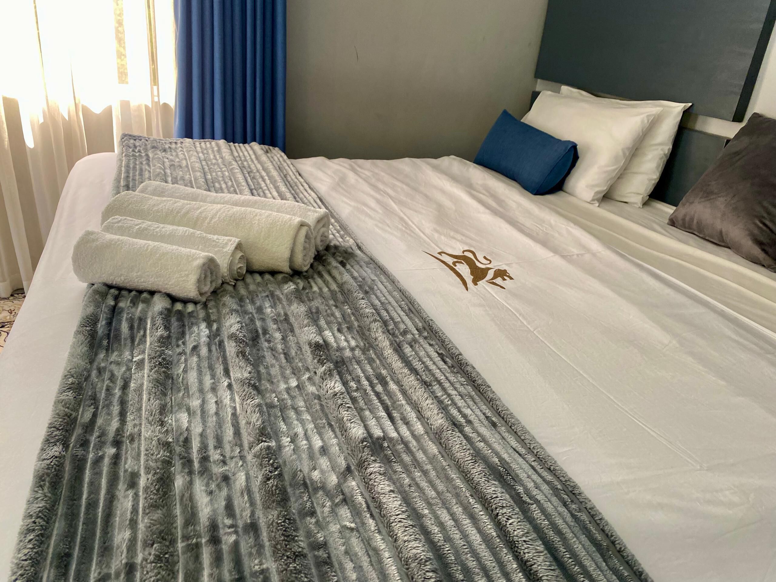 A neatly made bed with a grey and white striped blanket, a pair of rolled towels, and a decorative emblem.
