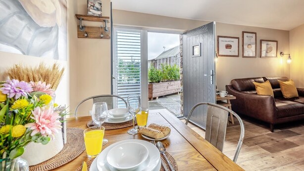 Ein Essbereich mit einem Holztisch, gedeckt für das Frühstück, einer braunen Ledercouch und Blick auf einen Garten durch eine offene Tür.