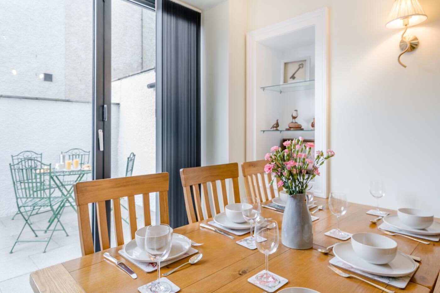 A dining table set for four with chairs, a vase of flowers, and a glass shelf with decorative items.