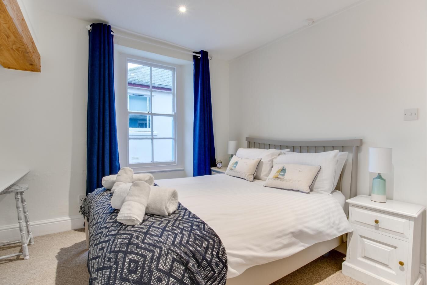 A neatly made bed with white linens and decorative pillows, a bedside table with a lamp, and a window with blue curtains.