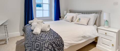 A neatly made bed with white linens and decorative pillows, a bedside table with a lamp, and a window with blue curtains.