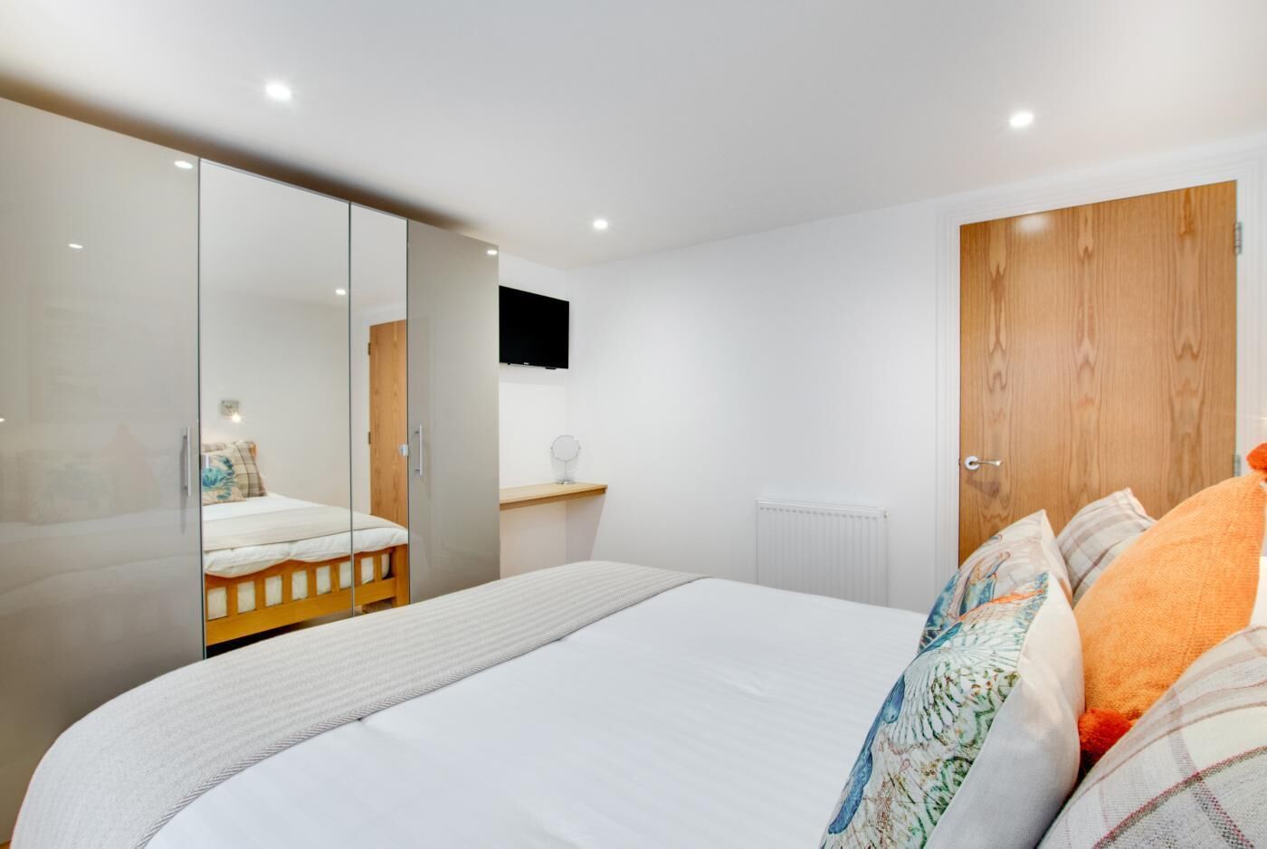 A modern bedroom with a large bed, a wooden wardrobe, a mirror, and a mounted TV.