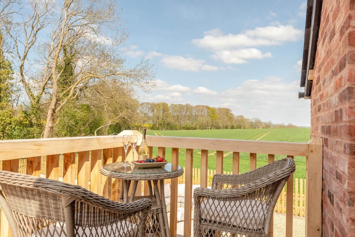 A wooden deck with wicker chairs and a table, overlooking a green field and trees.