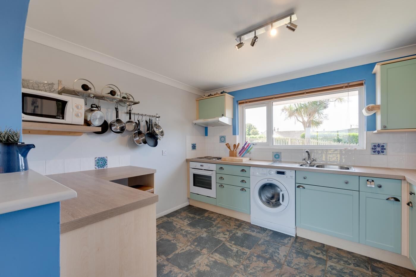 A modern kitchen with a built-in oven, microwave, and washing machine. There is a window providing natural light and a view of the outdoors.