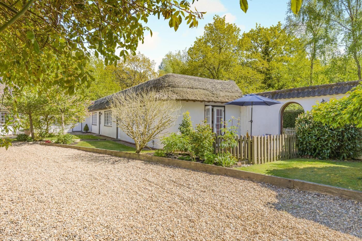 A thatched-roof cottage with a gravel driveway, surrounded by greenery and a wooden fence.