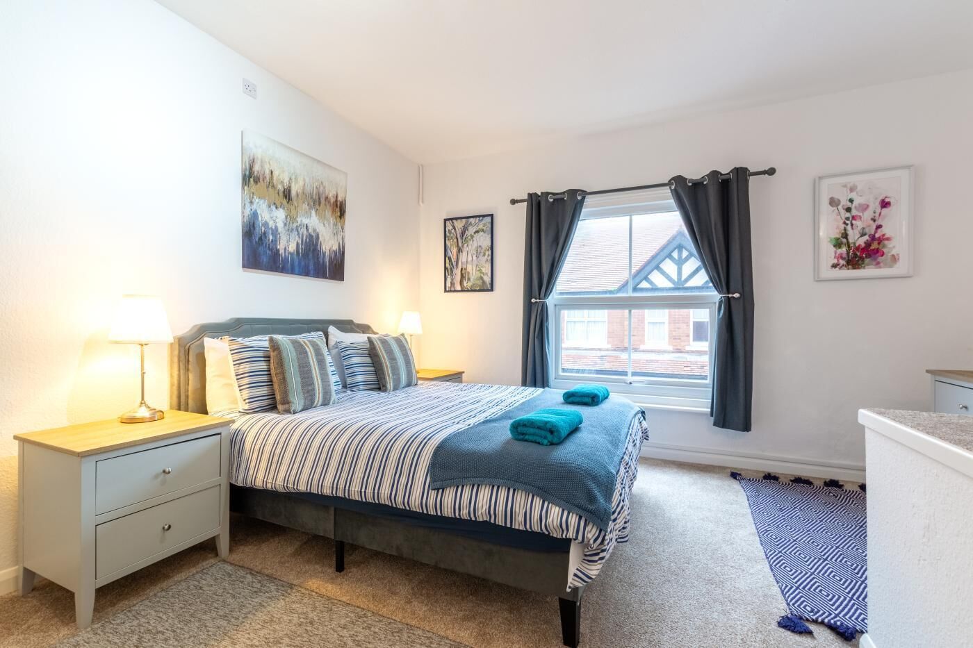 A bedroom with a bed, bedside tables, a window with curtains, and artwork on the walls.
