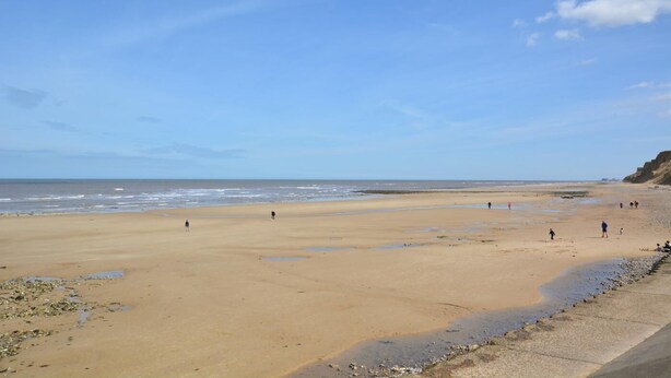 Ein weiter, sandiger Strand mit ruhiger See, einige Spaziergänger und ein Felsen am rechten Rand.