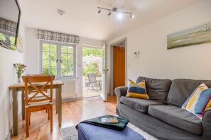A cozy living room with a grey sofa, wooden dining table, and a view of the garden through the door. - 1 Bed in West Runton (oc-1718) (West Runton)