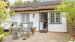 Ein weißes Cottage mit einem Strohdach, einer kleinen Terrasse mit Tisch und Stühlen und einem Garten mit Blumen und Pflanzen.