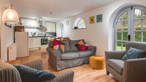 Een moderne woonkamer met een grijze bank, gestreepte fauteuils en een geel poef. In de achtergrond is een keuken zichtbaar met witte kasten en een eethoek.