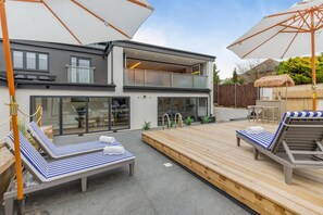 A modern two-story house with a patio area featuring sun loungers and umbrellas.