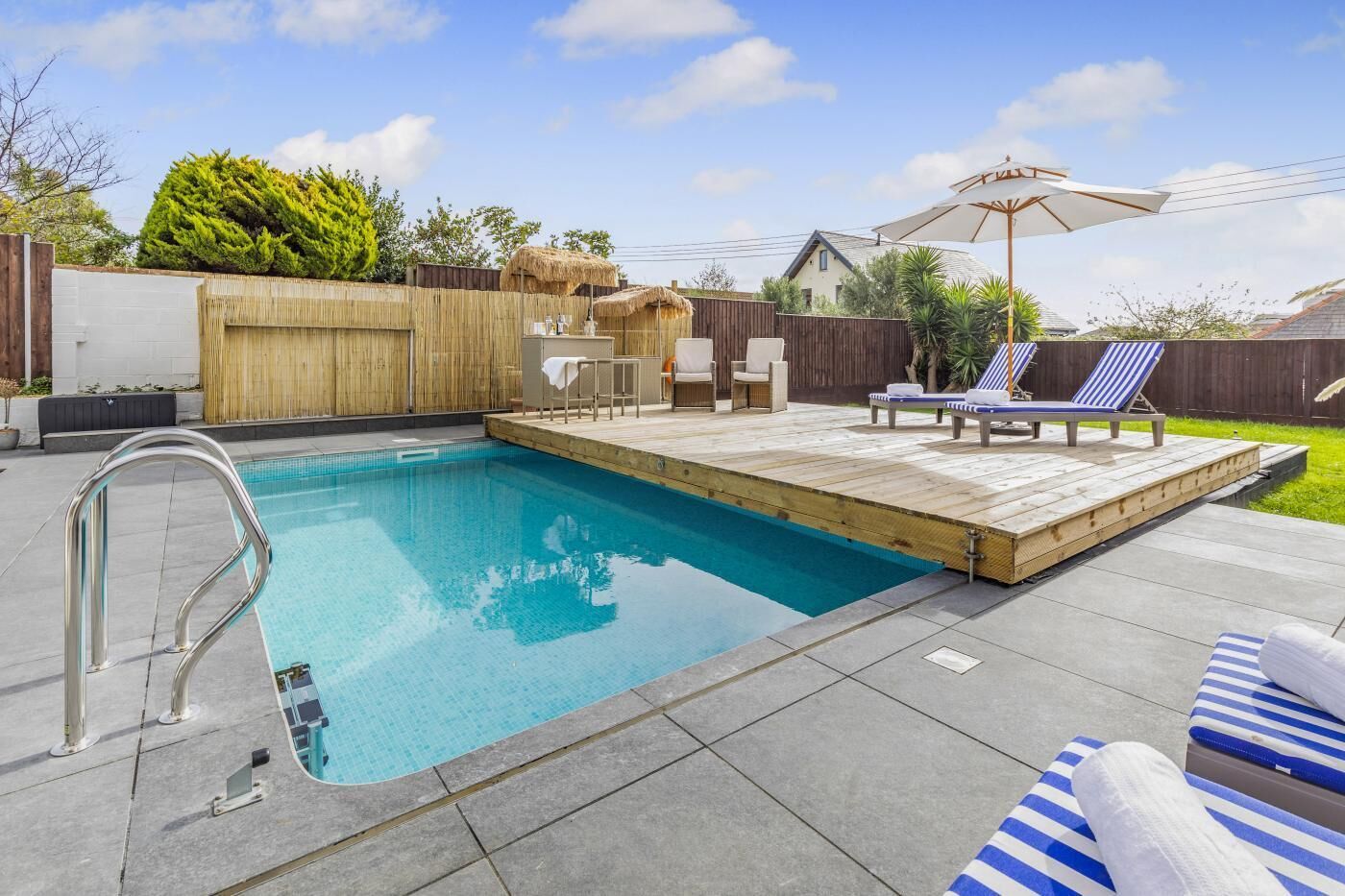 A rectangular swimming pool with a metal ladder on one side, surrounded by a wooden deck. There are lounge chairs and a table with chairs on the deck.