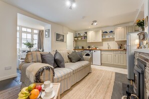 A cozy living room with a sofa, a dining table with fruit and cups, a kitchen with a washing machine, and a fireplace. - 2 Bed in Padstow (oc-p00572) (Padstow)