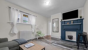 A modern living room with a sofa, a wooden coffee table, a hanging chair, a fireplace, and a TV mounted above it. - Furnished 2BR w/Parking – Mid-Term Stay Near Falls (Niagara Falls)