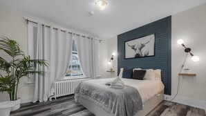 A modern bedroom with a bed, white curtains, a radiator, a potted plant, and a city view. - Furnished 2BR w/Parking – Mid-Term Stay Near Falls (Niagara Falls)