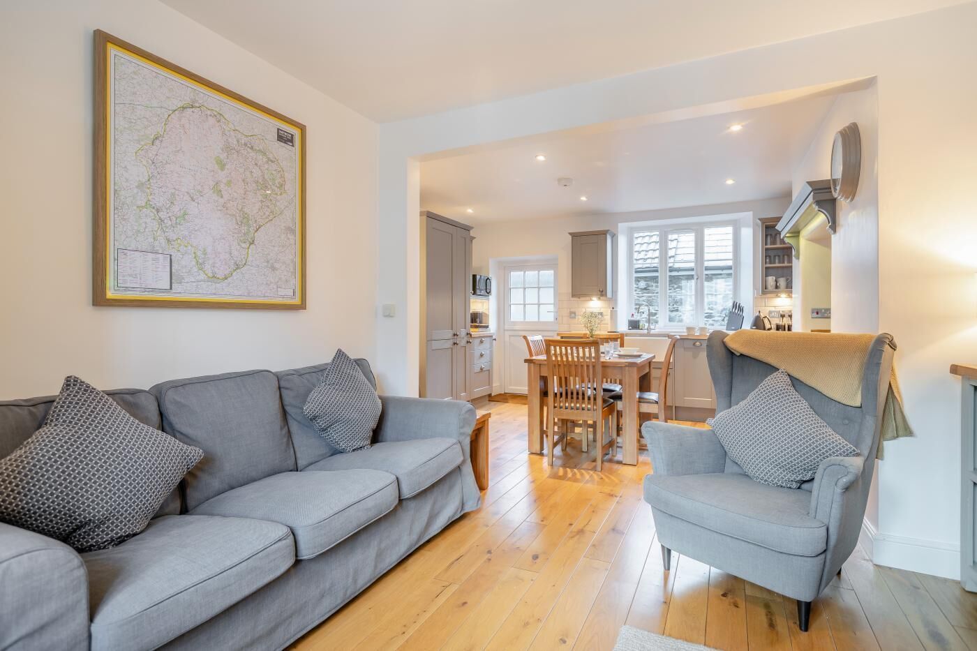 A spacious living room with a grey sofa and armchair, a dining area with wooden furniture, and a kitchen with white cabinets and a large window.