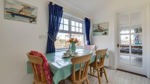 Un coin repas avec une table dressée pour deux personnes, des chaises en bois, un vase avec des fleurs et une fenêtre ornée de rideaux bleus.
