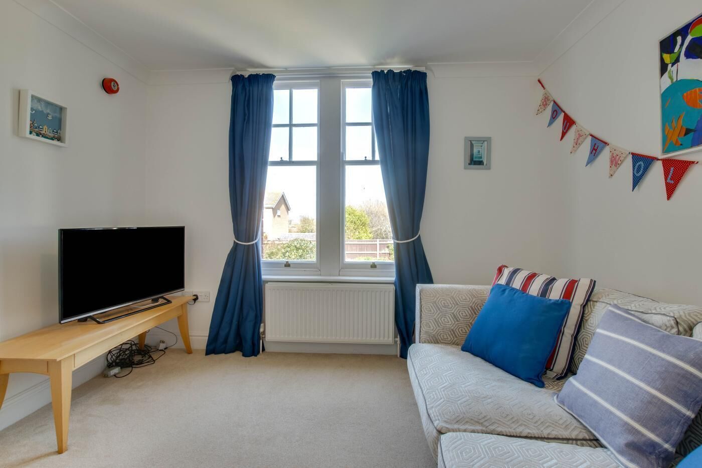 A living room with a television on a wooden stand, a sofa with patterned cushions, and a window with blue curtains.
