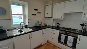 A compact kitchen with white cabinets, a black countertop, a built-in dishwasher, a gas stove, and a window with a view of the sea. - 2 Bed in Torquay (oc-9marco) (Torquay)