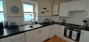 A compact kitchen with white cabinets, a black countertop, a built-in dishwasher, a gas stove, and a window with a view of the sea. - 2 Bed in Torquay (oc-9marco) (Torquay)