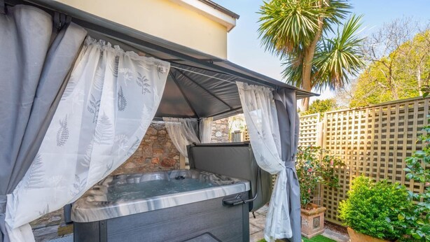 An outdoor hot tub with a stone wall backdrop, surrounded by a wooden fence and lush greenery. - 2 Bed in Torquay (oc-babbme) (Torquay)