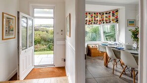 A dining area with a view of the garden, a table set for two, and a window with a colorful leaf-patterned curtain. - 4 Bed in Woolacombe (oc-h33441) (Woolacombe)