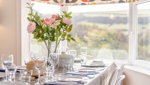A dining table set for four with a vase of pink roses and greenery, clear glassware, and a view of a landscape through a window. - 4 Bed in Woolacombe (oc-h33441) (Woolacombe)