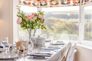 A dining table set for four with a vase of pink roses and greenery, clear glassware, and a view of a landscape through a window. - 4 Bed in Woolacombe (oc-h33441) (Woolacombe)