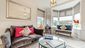 A living room with a brown leather sofa, a grey sofa, and a glass coffee table. There are two windows with views of greenery. - 4 Bed in Woolacombe (oc-h33441) (Woolacombe)