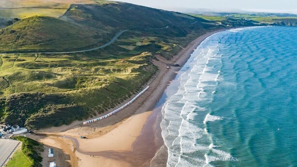 An aerial view of a coastal area with a beach, rolling hills, and a road curving along the coastline. - 4 Bed in Woolacombe (oc-h33441) (Woolacombe)