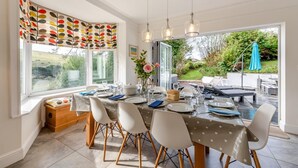 A dining area with a table set for four, large windows, and a view of the garden. - 4 Bed in Woolacombe (oc-h33441) (Woolacombe)