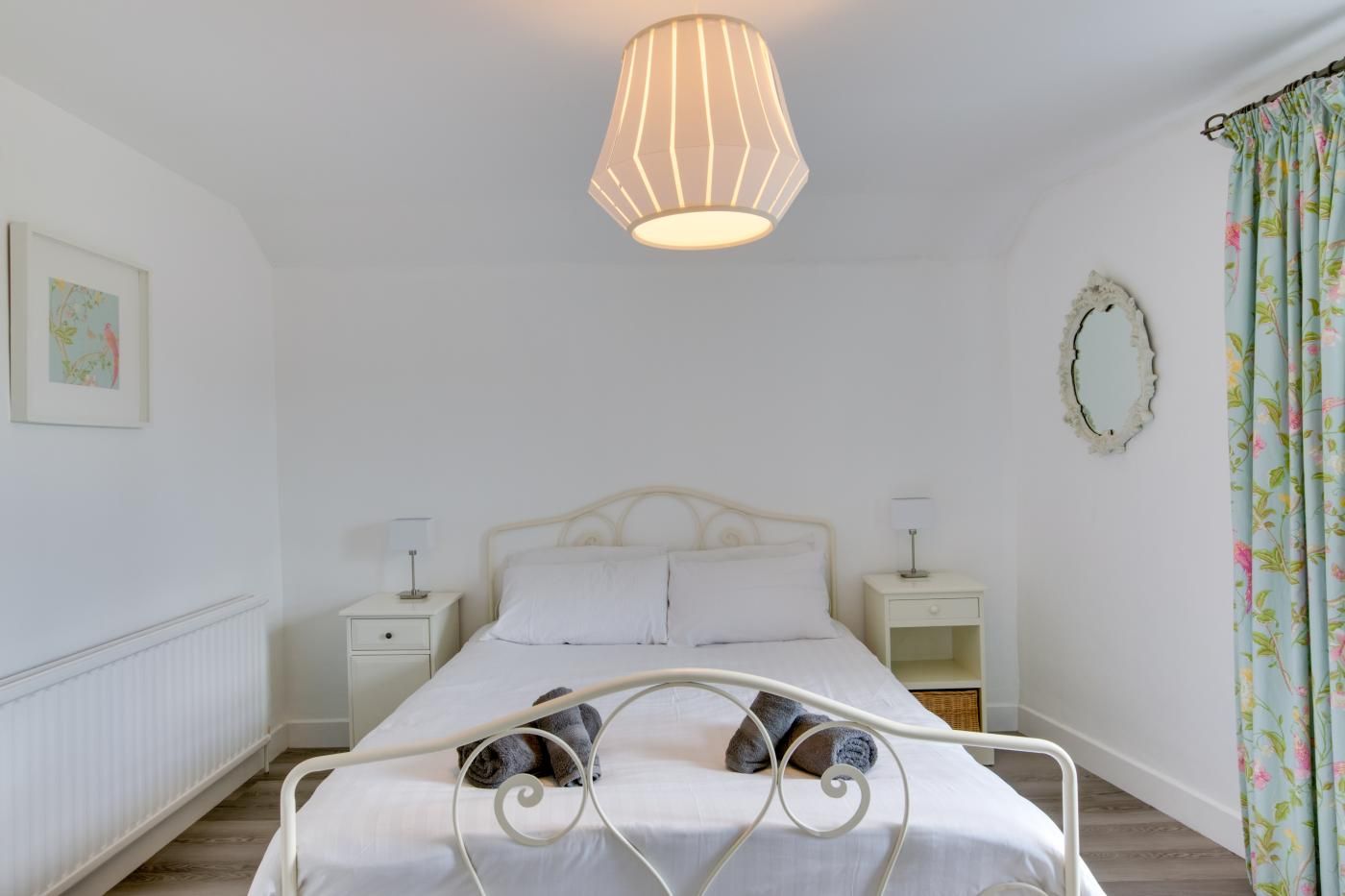 Ein Schlafzimmer mit einem Bett, zwei Nachttischen, einer Hängeleuchte, einem Heizkörper, einem gerahmten Bild, einem Spiegel und einem blumigen Vorhang.