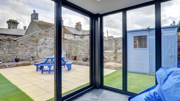 A modern room with large glass doors leading to a patio with a blue table and chairs, a stone wall, and a small shed. - 2 Bed in Beaumaris (oc-wai206) (Beaumaris)